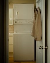 A narrow laundry space features a stacked white washer and dryer positioned against the wall. A beige shirt is hung on the back of the open door leading to the room. The walls and cabinetry are a soft, neutral color, complementing the appliances.