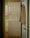 A narrow laundry space features a stacked white washer and dryer positioned against the wall. A beige shirt is hung on the back of the open door leading to the room. The walls and cabinetry are a soft, neutral color, complementing the appliances.