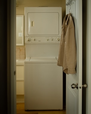 A narrow laundry space features a stacked white washer and dryer positioned against the wall. A beige shirt is hung on the back of the open door leading to the room. The walls and cabinetry are a soft, neutral color, complementing the appliances.
