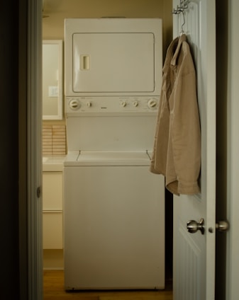 A narrow laundry space features a stacked white washer and dryer positioned against the wall. A beige shirt is hung on the back of the open door leading to the room. The walls and cabinetry are a soft, neutral color, complementing the appliances.