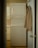 A narrow laundry space features a stacked white washer and dryer positioned against the wall. A beige shirt is hung on the back of the open door leading to the room. The walls and cabinetry are a soft, neutral color, complementing the appliances.