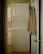A narrow laundry space features a stacked white washer and dryer positioned against the wall. A beige shirt is hung on the back of the open door leading to the room. The walls and cabinetry are a soft, neutral color, complementing the appliances.