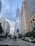 A towering skyscraper with reflective glass panels stands prominently in a cityscape. Several other tall buildings and office structures surround it. The sky is partly cloudy, and a busy street with vehicles, including a delivery truck and several cars, is visible below.