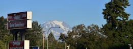 A tall mountain with snow-covered peaks is visible in the background. In the foreground, there's a sign for a grocery or convenience store with the words 'Super Saver Foods' and 'Open 24 Hours'. The scene includes trees and utility lines, suggesting a suburban setting on a clear day.