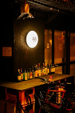 A warmly lit bar with a variety of liquor bottles arranged neatly on a wooden counter. A bicycle with a basket is parked in front, adding an urban touch. A glowing round sign with Japanese text is visible, contributing to a cozy ambiance.