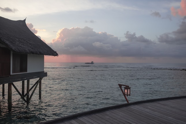 A tranquil ocean scene with a thatched-roofed overwater bungalow on stilts to the left, a soft sunset casting hues of pink and orange across the sky, reflected on the calm water. A distant boat is visible near the horizon, and a wooden deck with a lantern leads into the scene.