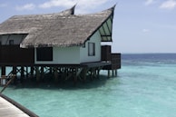 A thatched roof villa built on stilts over clear turquoise water. The villa has a wooden deck and traditional architecture. The ocean surrounding the structure is calm and vast, extending to the horizon under a partly cloudy sky.