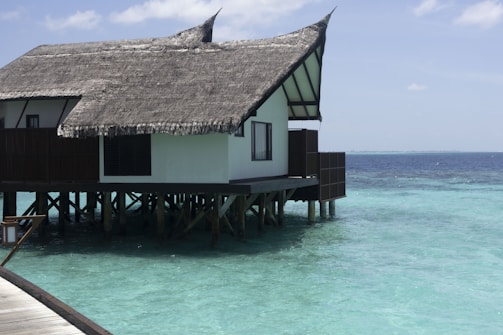A thatched roof villa built on stilts over clear turquoise water. The villa has a wooden deck and traditional architecture. The ocean surrounding the structure is calm and vast, extending to the horizon under a partly cloudy sky.