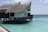 A thatched roof villa built on stilts over clear turquoise water. The villa has a wooden deck and traditional architecture. The ocean surrounding the structure is calm and vast, extending to the horizon under a partly cloudy sky.