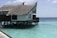 A thatched roof villa built on stilts over clear turquoise water. The villa has a wooden deck and traditional architecture. The ocean surrounding the structure is calm and vast, extending to the horizon under a partly cloudy sky.