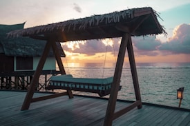 A wooden deck overlooking the ocean with a large swing bed under a thatched roof. The sun is setting in the background, casting a warm glow across the sky and water. Clouds are scattered in the sky, enhancing the tranquil and serene atmosphere.