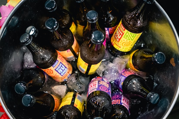 A bucket filled with ice and several brown glass bottles, each adorned with colorful labels featuring vibrant geometric designs and text. The bottles are standing upright and are partially submerged in the melting ice.
