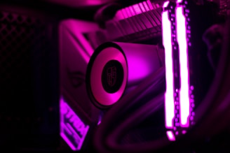 Close-up view of a computer's internal components illuminated by bright magenta lighting. The image shows the cooling system, including a circular component with a logo, and prominently lit RAM sticks. Tubes and cables are also visible, lit up by the intense magenta glow.