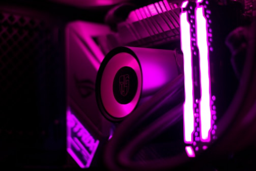 Close-up view of a computer's internal components illuminated by bright magenta lighting. The image shows the cooling system, including a circular component with a logo, and prominently lit RAM sticks. Tubes and cables are also visible, lit up by the intense magenta glow.