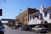 A street scene at the Fort Worth Stock Yards, featuring historical brick buildings and a sign overhead indicating the location. On the right, there is a white building with a model horse on the balcony and signage for a western store. A line of cars and trucks is parked along the cobblestone street, and there is an American flag flying in the background.
