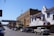A street scene at the Fort Worth Stock Yards, featuring historical brick buildings and a sign overhead indicating the location. On the right, there is a white building with a model horse on the balcony and signage for a western store. A line of cars and trucks is parked along the cobblestone street, and there is an American flag flying in the background.