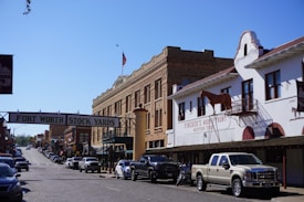 A street scene at the Fort Worth Stock Yards, featuring historical brick buildings and a sign overhead indicating the location. On the right, there is a white building with a model horse on the balcony and signage for a western store. A line of cars and trucks is parked along the cobblestone street, and there is an American flag flying in the background.