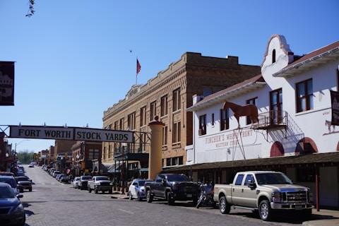 A street scene at the Fort Worth Stock Yards, featuring historical brick buildings and a sign overhead indicating the location. On the right, there is a white building with a model horse on the balcony and signage for a western store. A line of cars and trucks is parked along the cobblestone street, and there is an American flag flying in the background.