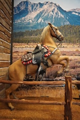 A horse, appearing lifelike, is rearing up on its hind legs. The horse is saddled and bridled, suggesting it is ready for riding. Behind the horse, a large mural depicts a breathtaking mountain landscape with clear blue skies, forested hills, and grassy plains.
