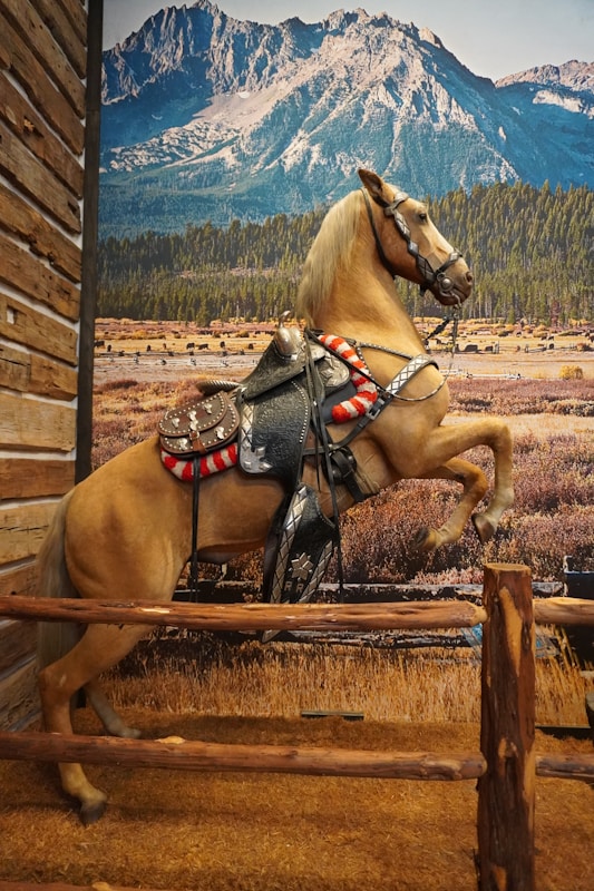 A horse, appearing lifelike, is rearing up on its hind legs. The horse is saddled and bridled, suggesting it is ready for riding. Behind the horse, a large mural depicts a breathtaking mountain landscape with clear blue skies, forested hills, and grassy plains.