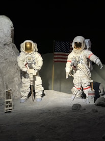 Two astronauts in white space suits are standing on a rocky, barren surface. One astronaut is holding equipment, and the other is gesturing with an arm extended. In the background, an American flag is planted in the ground. The setting appears to be a representation of the lunar landscape with gray ground and dark, empty space surrounding them.