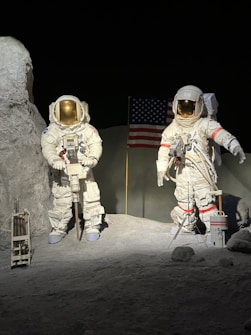 Two astronauts in white space suits are standing on a rocky, barren surface. One astronaut is holding equipment, and the other is gesturing with an arm extended. In the background, an American flag is planted in the ground. The setting appears to be a representation of the lunar landscape with gray ground and dark, empty space surrounding them.