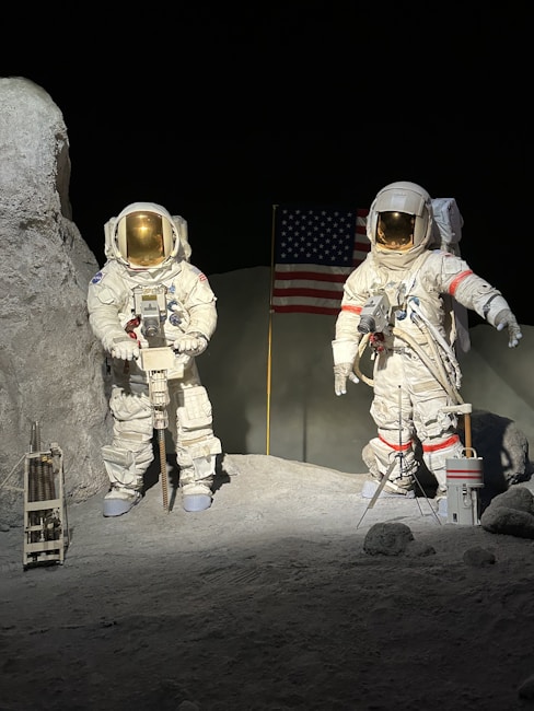 Two astronauts in white space suits are standing on a rocky, barren surface. One astronaut is holding equipment, and the other is gesturing with an arm extended. In the background, an American flag is planted in the ground. The setting appears to be a representation of the lunar landscape with gray ground and dark, empty space surrounding them.