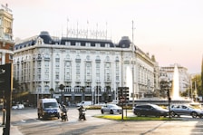 A grand, historic hotel building featuring ornate architectural details with a large sign reading 'PALACE HOTEL' on the roof. The area in front includes a busy traffic circle with cars, motorcycles, and an emergency vehicle. There are also two large fountains, manicured landscaping, and surrounding buildings.