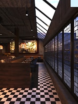 An interior setting with a modern dining area featuring checkered flooring and a row of windows with a geometric design. Warm lighting from hanging lamps creates a cozy ambiance, while a mural depicting foliage adds an artistic touch to the wall. Booth seating is arranged for dining, with tables set up and ready for service.