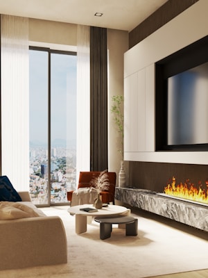 A modern living room with a large window offering a cityscape view. The room features a cozy beige sofa, a rust-colored armchair, and a set of stylish round coffee tables. A sleek fireplace with a marble hearth adds warmth. The design includes sheer curtains, a minimalist plant, and a large flat-screen TV mounted above the fireplace.