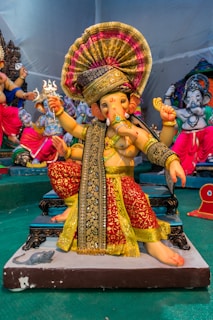 A colorful and detailed statue of a deity, intricately dressed in vibrant traditional clothing. The figure has multiple arms holding various symbolic items and is adorned with ornate decorations including a headdress and jewelry. Other similar statues are visible in the background.