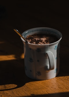 A ceramic mug with a floral pattern holds a heap of cocoa powder, and a spoon is partially submerged in the powder. The mug sits on a wooden surface, with soft light creating shadows and highlighting the texture of the wood and the design on the mug.