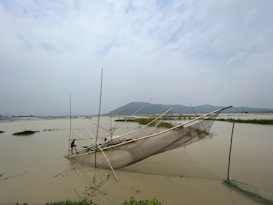A large fishing net is set up in a calm body of water, with a person operating it. The scene is peaceful, with distant hills and a cloudy sky