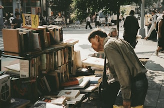 A man leans over a makeshift bookstand piled with various books on a busy city street corner. Pedestrians are seen walking by in the background, and there are trees providing some shade. The atmosphere suggests the vibrancy and hustle of urban life.