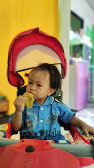 A young child wearing a blue shirt is sitting in a stroller with a red canopy, eating a piece of food. The background features a yellow wall and various colorful elements, indicating an indoor setting.