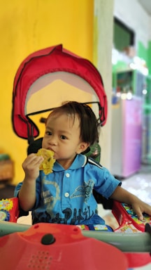 A young child wearing a blue shirt is sitting in a stroller with a red canopy, eating a piece of food. The background features a yellow wall and various colorful elements, indicating an indoor setting.