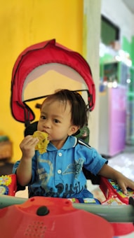 A young child wearing a blue shirt is sitting in a stroller with a red canopy, eating a piece of food. The background features a yellow wall and various colorful elements, indicating an indoor setting.