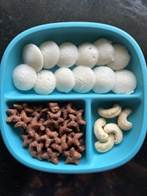 A blue divided plate contains three sections of snacks: white steamed rice cakes, brown star-shaped cereal pieces, and a few cashew nuts.