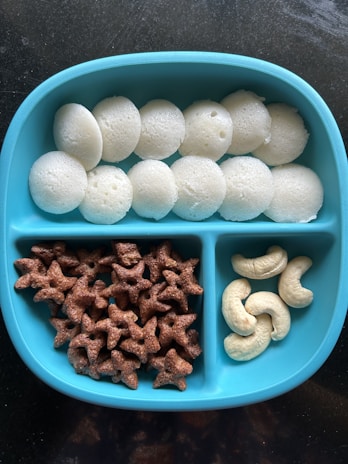 A blue divided plate contains three sections of snacks: white steamed rice cakes, brown star-shaped cereal pieces, and a few cashew nuts.
