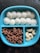 A blue divided plate contains three sections of snacks: white steamed rice cakes, brown star-shaped cereal pieces, and a few cashew nuts.