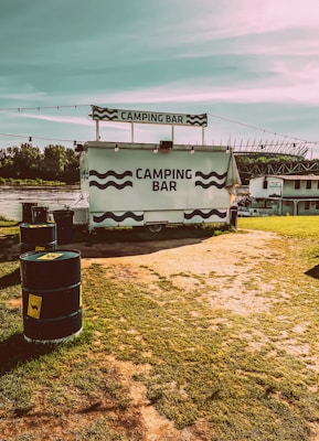 A mobile bar labeled 'Camping Bar' is set up on a grassy area near a body of water. The bar is positioned with a small overhead sign and surrounded by several black barrels with yellow labels. A shaded walkway and some structures can be seen in the distance under a clear blue sky.