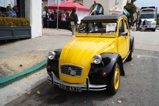 A vintage yellow and black car is parked on the street with a 'for sale' sign on its windshield. The car has a classic design with round headlights and a curved roof. In the background, a group of people is gathered near a red tent and buildings with colorful awnings. A large truck is visible further up the street.