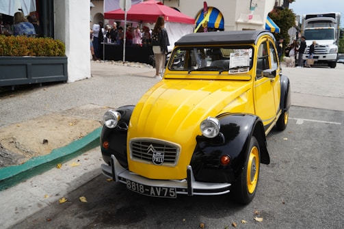 A vintage yellow and black car is parked on the street with a 'for sale' sign on its windshield. The car has a classic design with round headlights and a curved roof. In the background, a group of people is gathered near a red tent and buildings with colorful awnings. A large truck is visible further up the street.