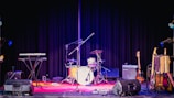 A stage setup featuring a variety of musical instruments including a drum set, electric guitar, keyboard, and amplifiers. The background is a black curtain and the stage is illuminated with colorful stage lights.