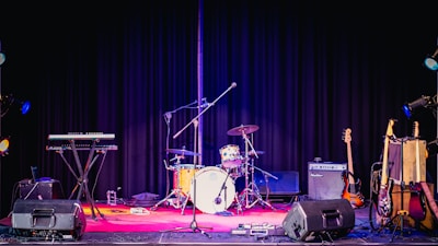 A stage setup featuring a variety of musical instruments including a drum set, electric guitar, keyboard, and amplifiers. The background is a black curtain and the stage is illuminated with colorful stage lights.