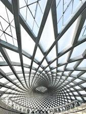 A spiraling architectural ceiling design consisting of interlocking beams and glass panels, forming a geometric pattern that creates a sense of depth and movement. The light filters through the glass, casting subtle shadows.