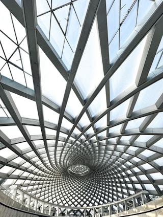 A spiraling architectural ceiling design consisting of interlocking beams and glass panels, forming a geometric pattern that creates a sense of depth and movement. The light filters through the glass, casting subtle shadows.