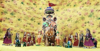 A collection of colorful, traditional Rajasthani puppets is displayed against a floral yellow background. The central figure is an elaborately decorated horse with a rider, surrounded by smaller human and animal figures, all adorned with traditional Rajasthani attire and accessories.