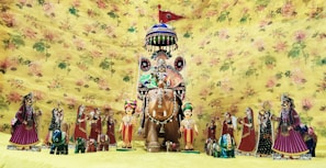 A collection of colorful, traditional Rajasthani puppets is displayed against a floral yellow background. The central figure is an elaborately decorated horse with a rider, surrounded by smaller human and animal figures, all adorned with traditional Rajasthani attire and accessories.