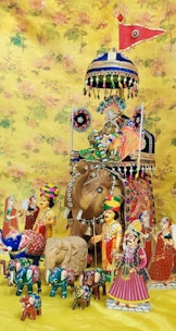A colorful arrangement of traditional Indian dolls and figurines is displayed against a floral yellow backdrop. The centerpiece features a large wooden elephant adorned with intricate designs, topped with a vibrant umbrella and a red flag. Surrounding the elephant are various figures, including human characters dressed in traditional attire, and smaller elephants painted in bright colors.